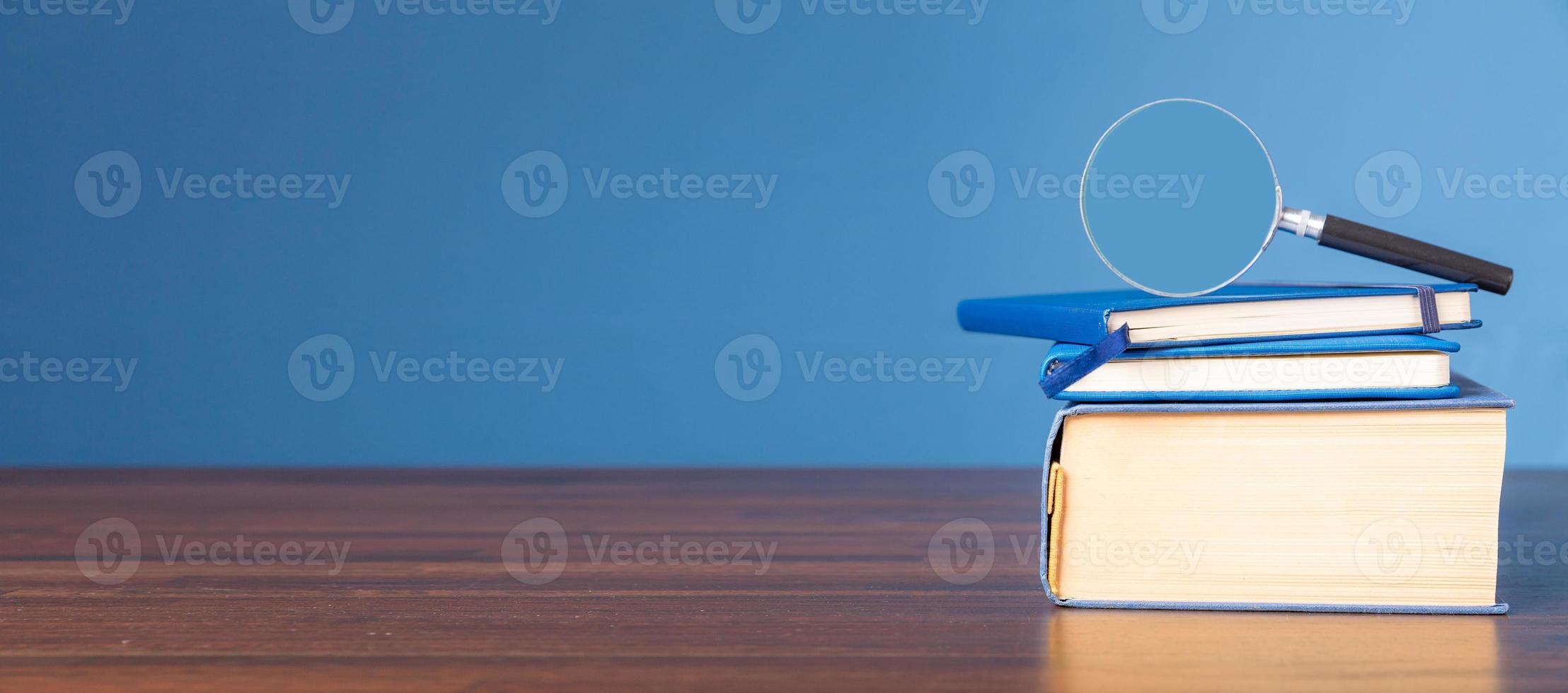 stack bok med förstoringsglas på trä skrivbord i informationsbibliotek av skolan eller universitetet, koncept för utbildning, läsning, studier, kopiera utrymme på blå bakgrund. foto