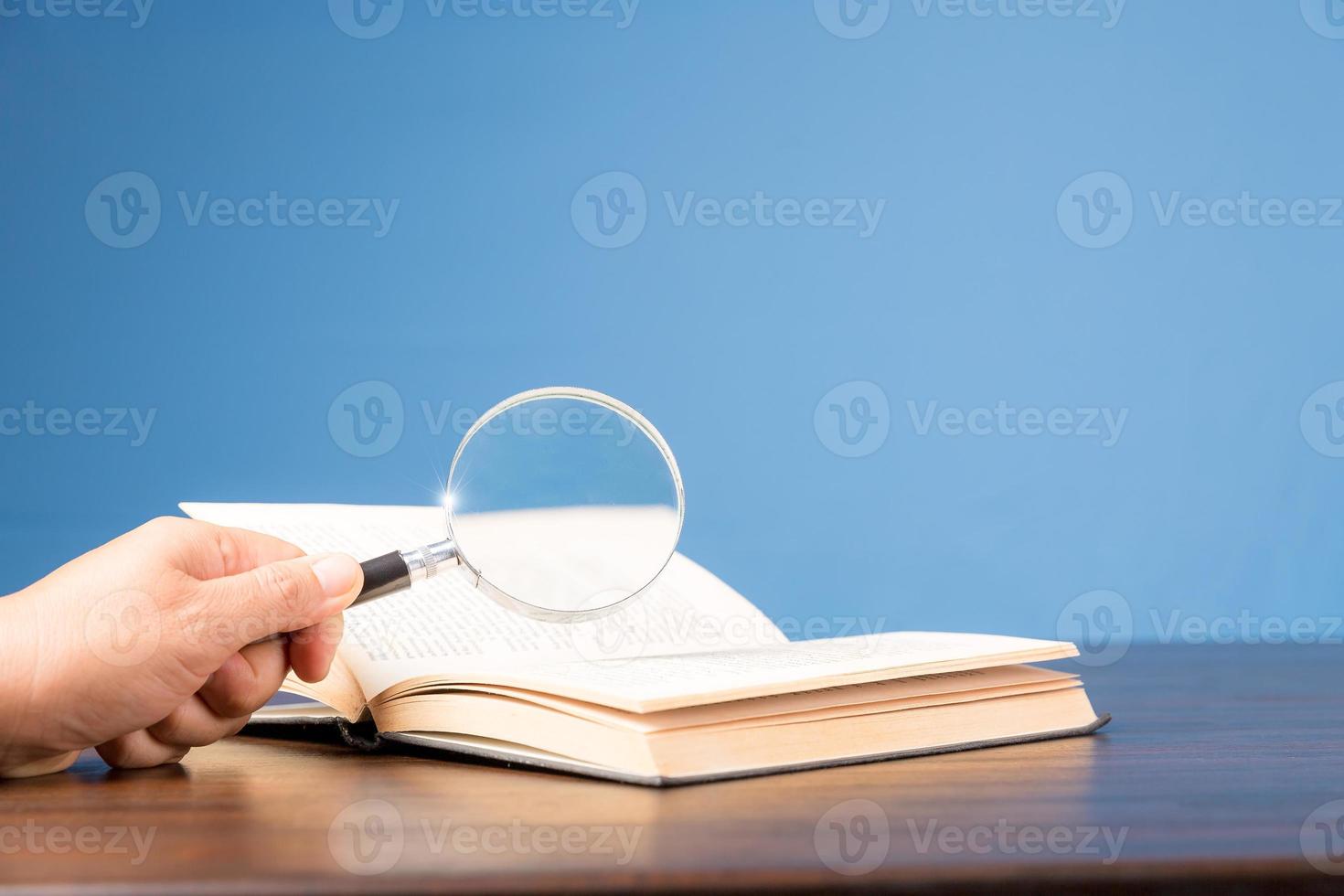 öppen bok med förstoringsglas på träskrivbord i informationsbibliotek för skolan eller universitetet, koncept för utbildning, läsning, studier, kopieringsutrymme och blå bakgrund. foto