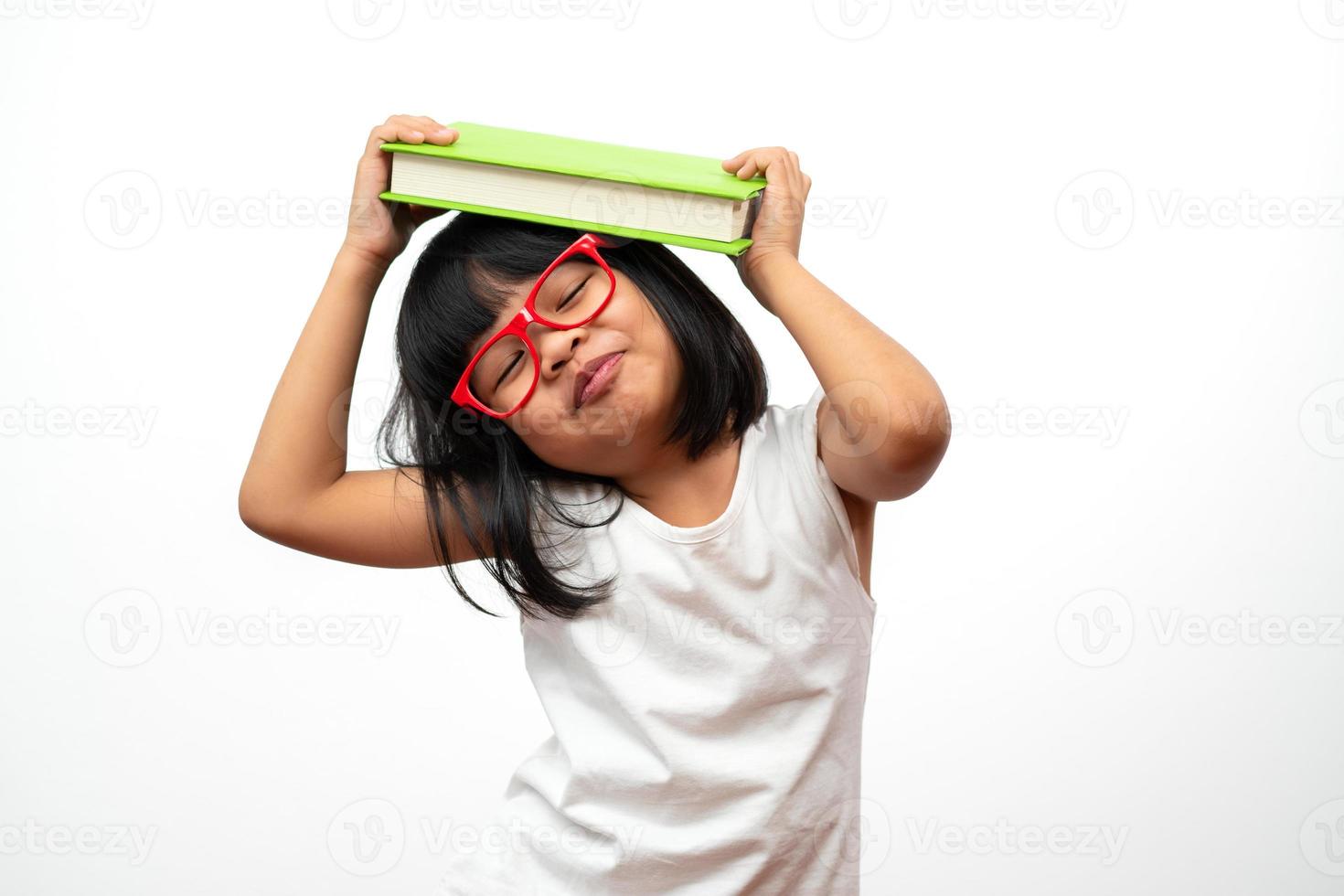 rolig och glad asiatisk liten förskoleflicka som bär röda glasögon som håller en grön bok på huvudet, på vit isolerad bakgrund. begreppet skolbarn och utbildning i grundskolan och förskolan foto