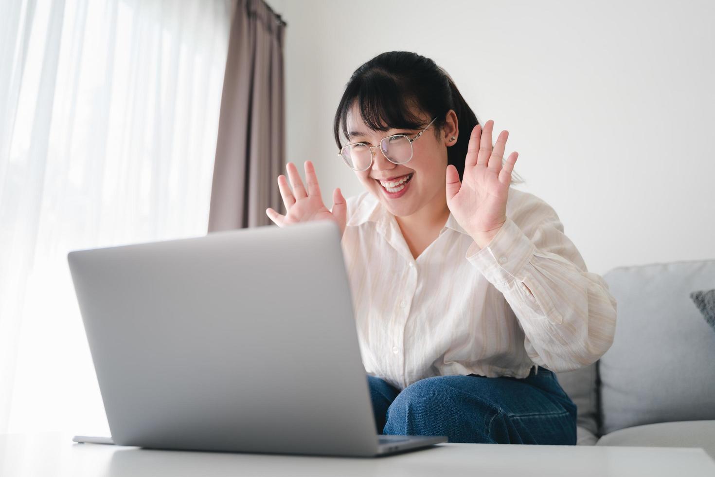 ung asiatisk kvinna som använder bärbar dator för online videokonferenssamtal viftande hand gör hej gest på soffan i vardagsrummet. foto