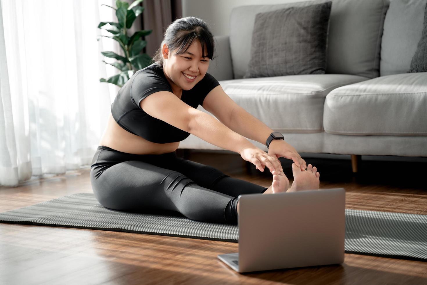 asiatisk knubbig kvinna sitter på golvet i vardagsrummet öva online yogalektion med datorn. kvinna med meditera träningsklass på den bärbara datorn. foto