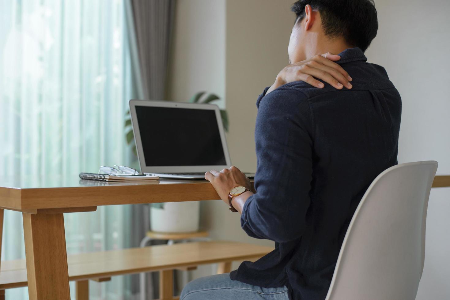 en ung man sitter vid sitt skrivbord hemma med hjälp av en dator. han lägger händerna på nacken och axlarna när han känner smärta i musklerna av att sitta länge. foto