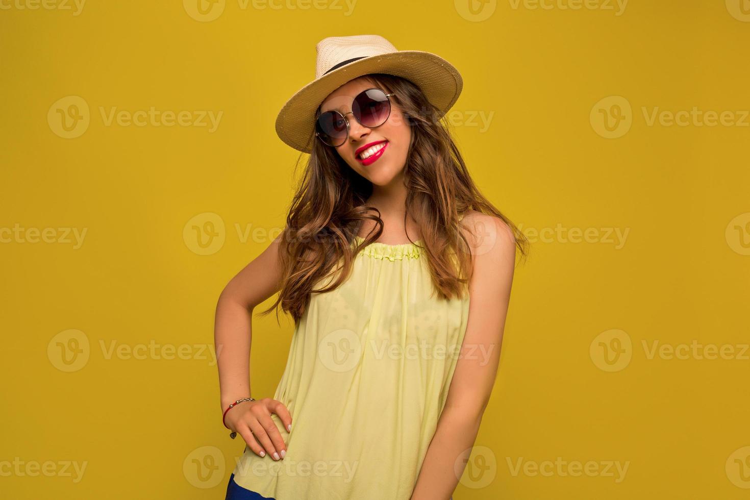 underbar brunett tjej i solglasögon och hatt poserar på gul bakgrund med ett glatt leende. skrattande kvinnlig modell i elegant klänning i studio foto