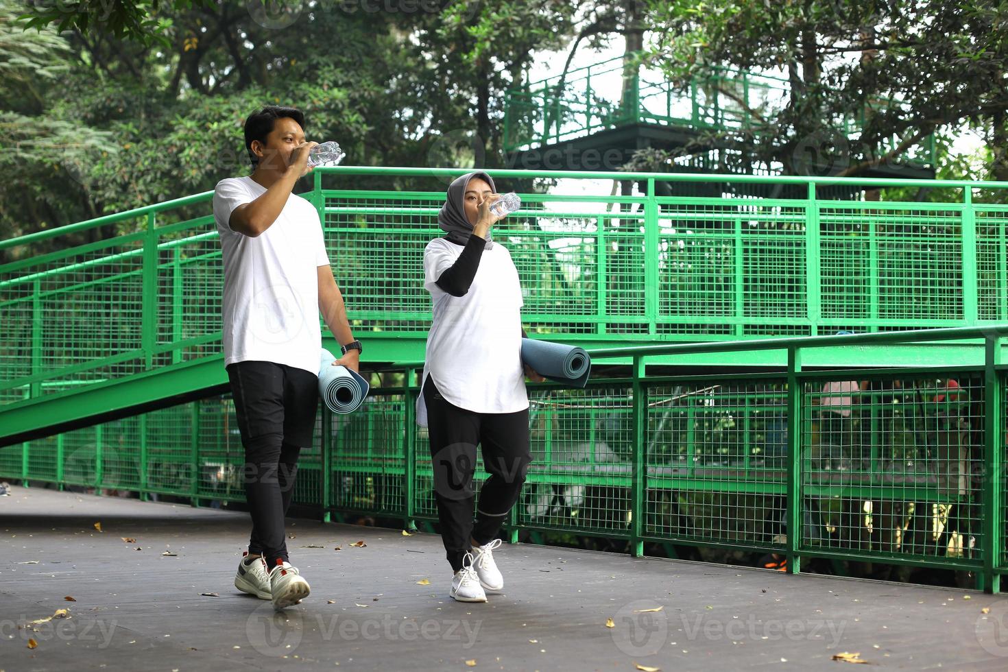 Ungt asiatiskt par dricker vatten efter träningspass medan de går och håller i madrassen efter träningspass i parken. foto