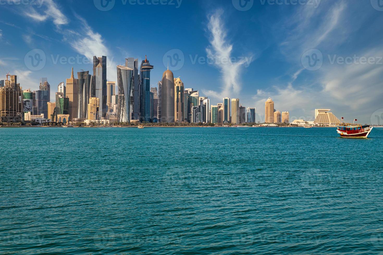 doha qatar skyline dagsljusvy med moln på himlen som visar skyskrapor och dhow med qatars flagga i bakgrunden och arabiska viken i förgrunden foto