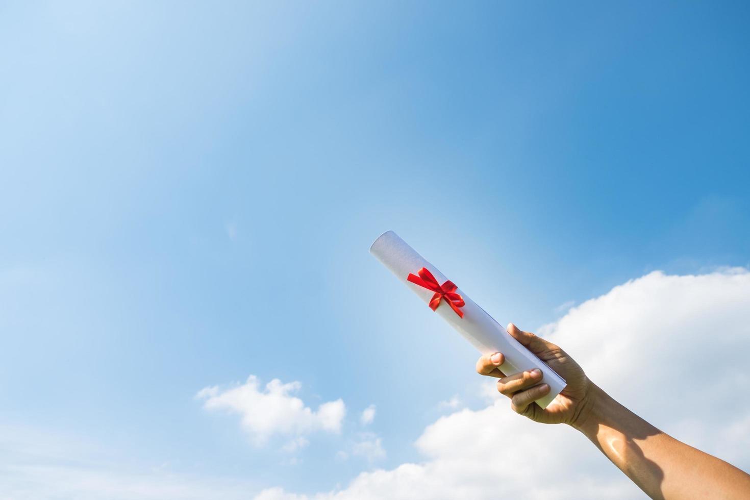 hand som håller examen certifikat rulle under solskenet, blå himmel bakgrund. foto