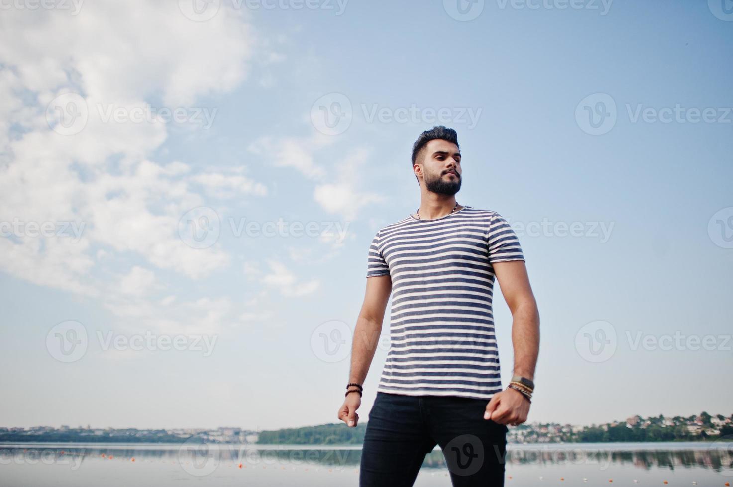 stilig lång arabisk skägg man modell på avskalad skjorta poserade utomhus mot sjön och himlen. moderiktig arabkille. foto