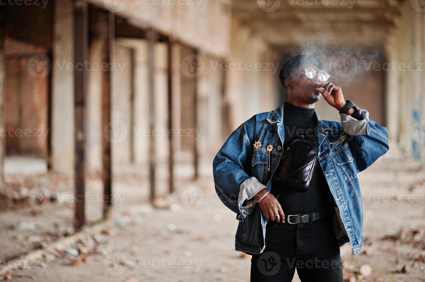 afroamerikansk man i jeansjacka, basker och glasögon, röker cigarr på övergiven fabrik. foto