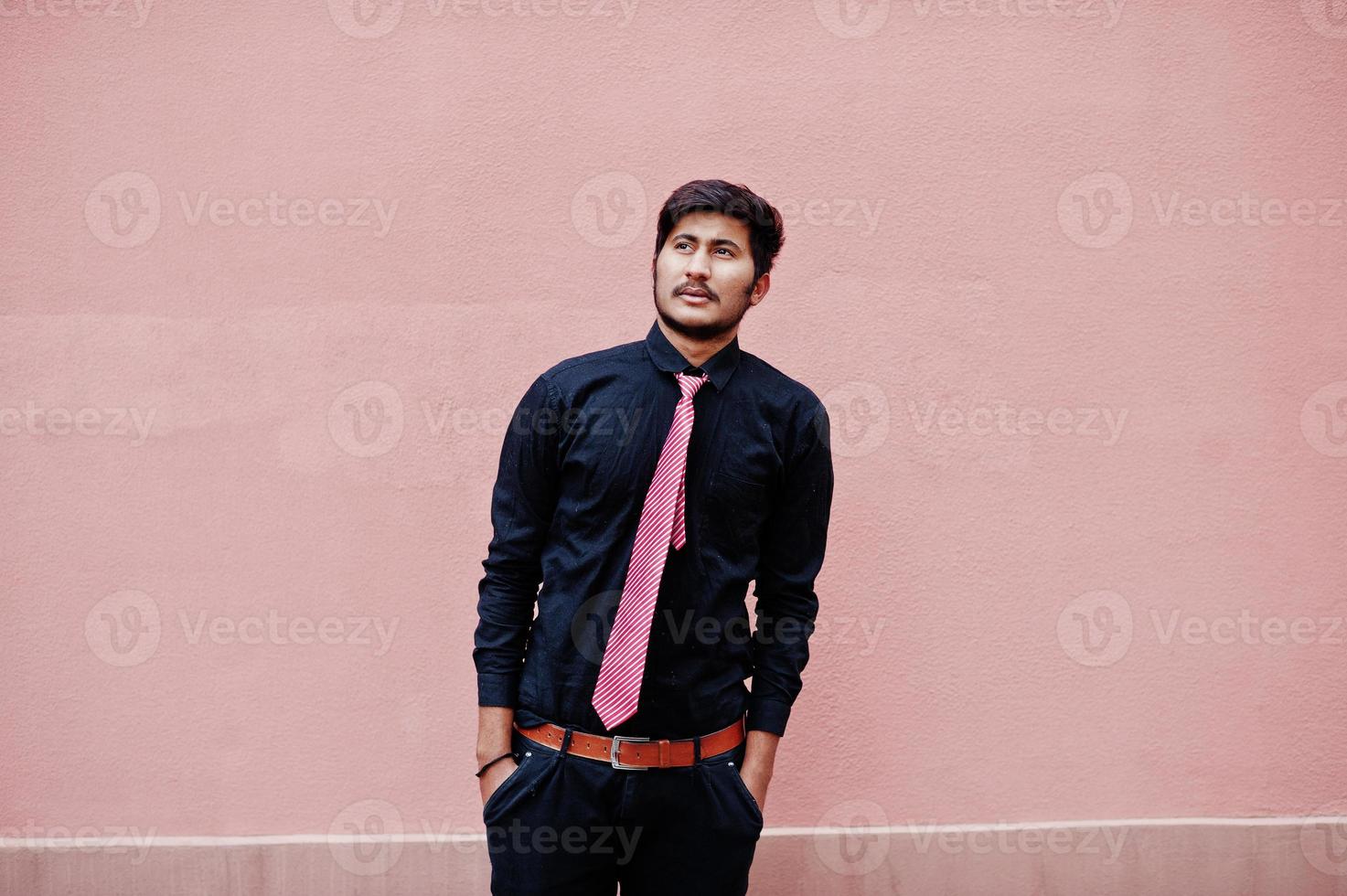 ung indisk man på svart skjorta och slips poserade mot den rosa väggen. foto