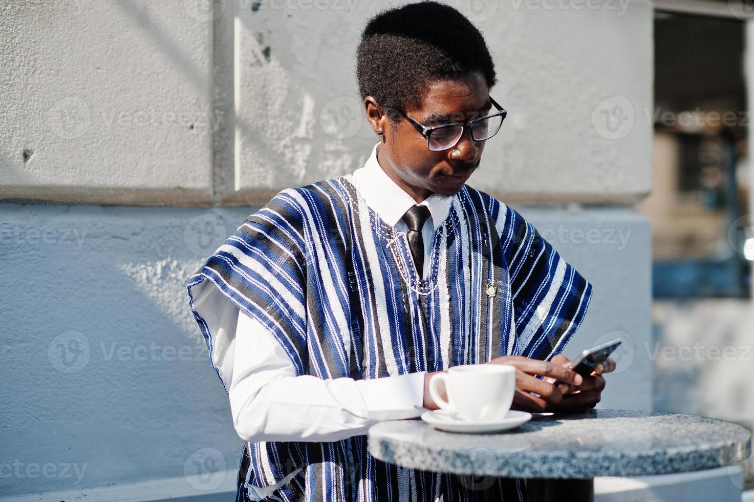 afrikansk man i traditionella kläder och glasögon på utomhuscafé, dricker kaffe och tittar på dagliga nyheter på telefonen. foto
