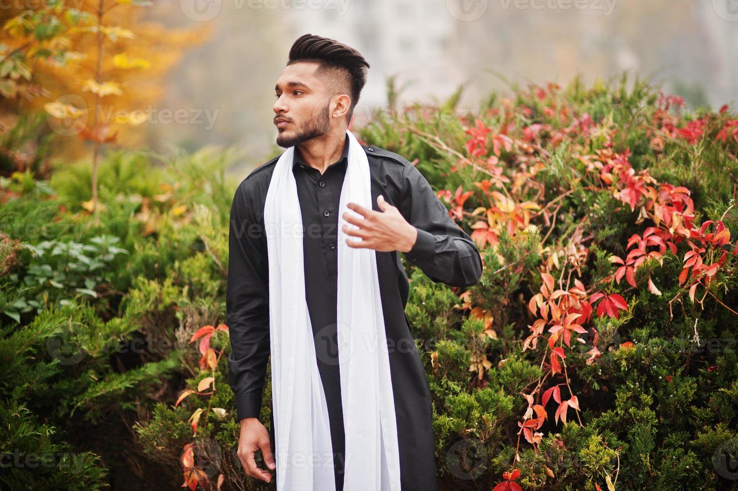 indisk snygg man i svarta traditionella kläder med vit halsduk poserade utomhus mot gula höstlöv träd. foto