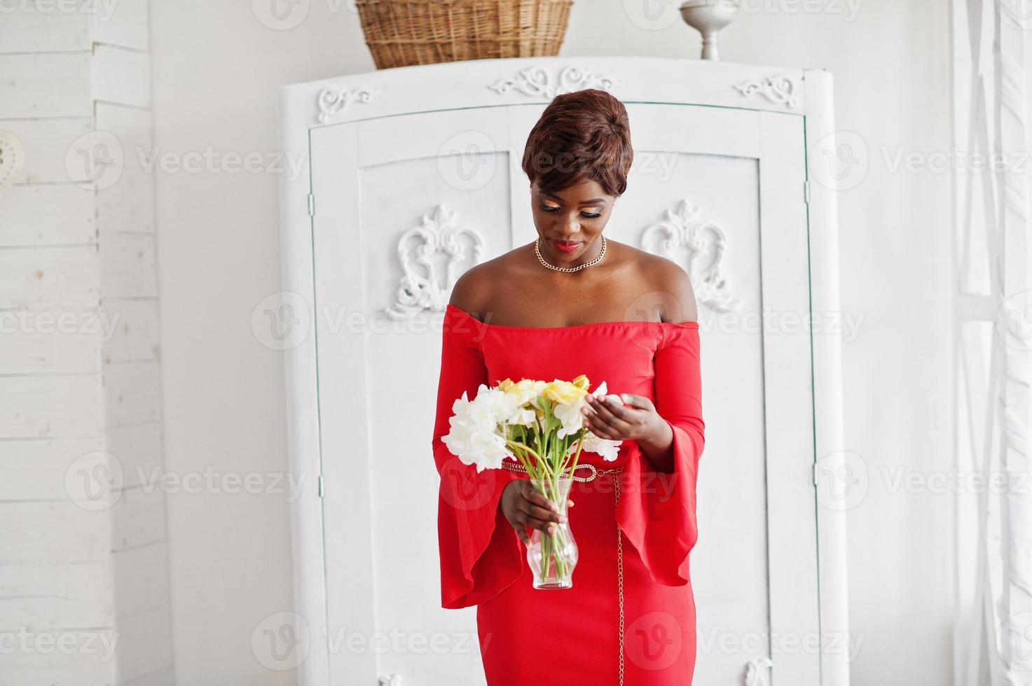 mode afrikansk amerikansk modell i röd skönhetsklänning, sexig kvinna poserar aftonklänning som håller blommor i vit vintagerum. foto