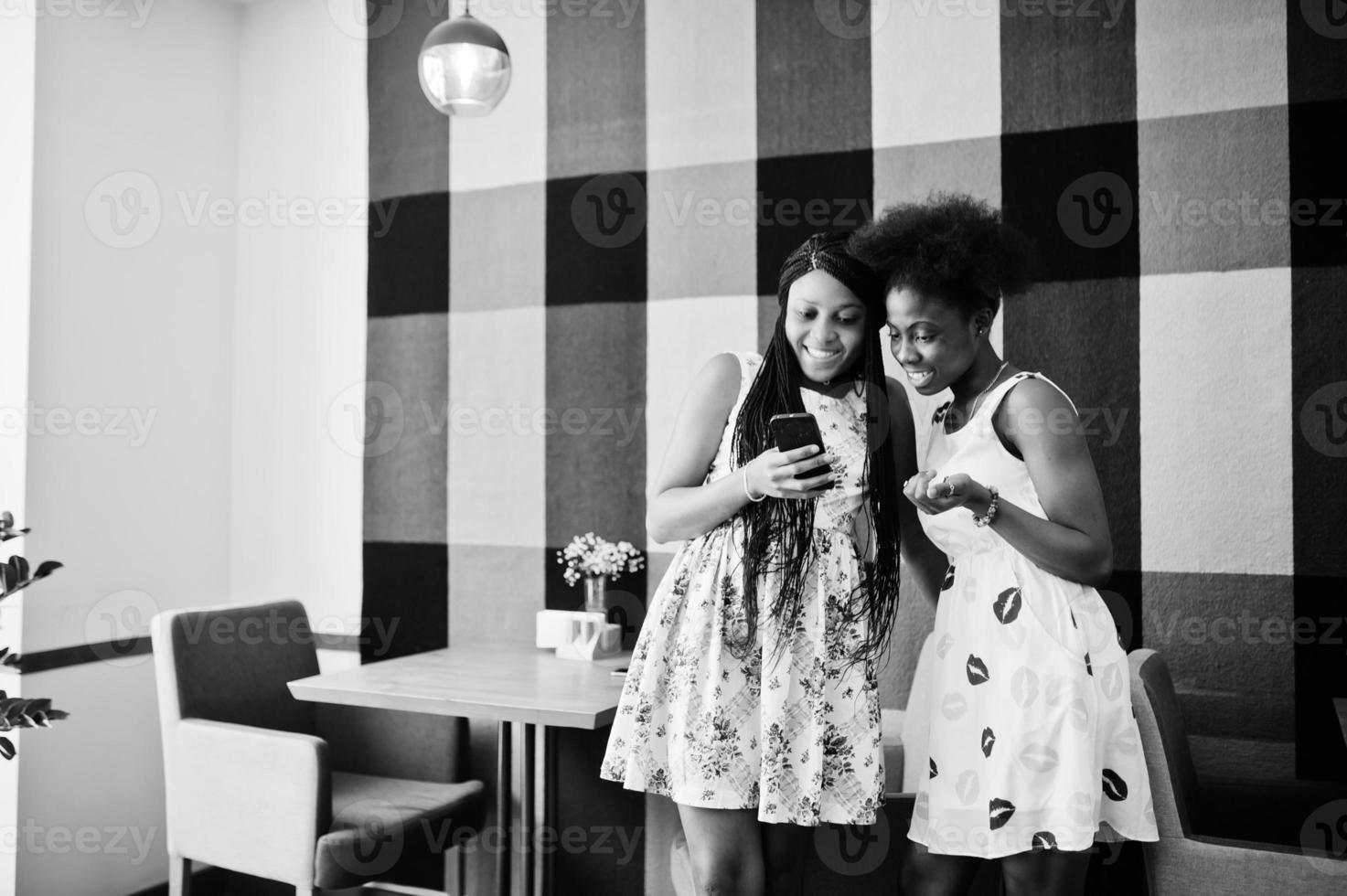 två glada svarta afrikanska flickvänner på sommarklänningar tittar på mobiltelefon på café. foto