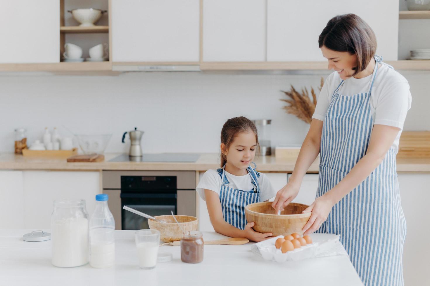 lilla dotter och mamma lagar kakor tillsammans hemma, klädda i förkläden, står mot köksinredningen, lagar söt måltid tillsammans, njuter av att spendera tid hemma. bakning, familjekoncept foto