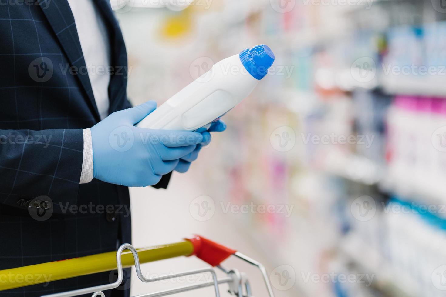 man väljer tvättmedel i hushållsbutik, håller flaska med flytande pulver, bär medicinska handskar för att skydda mot coronavirus, läser produktinformation. handlar under karantän foto