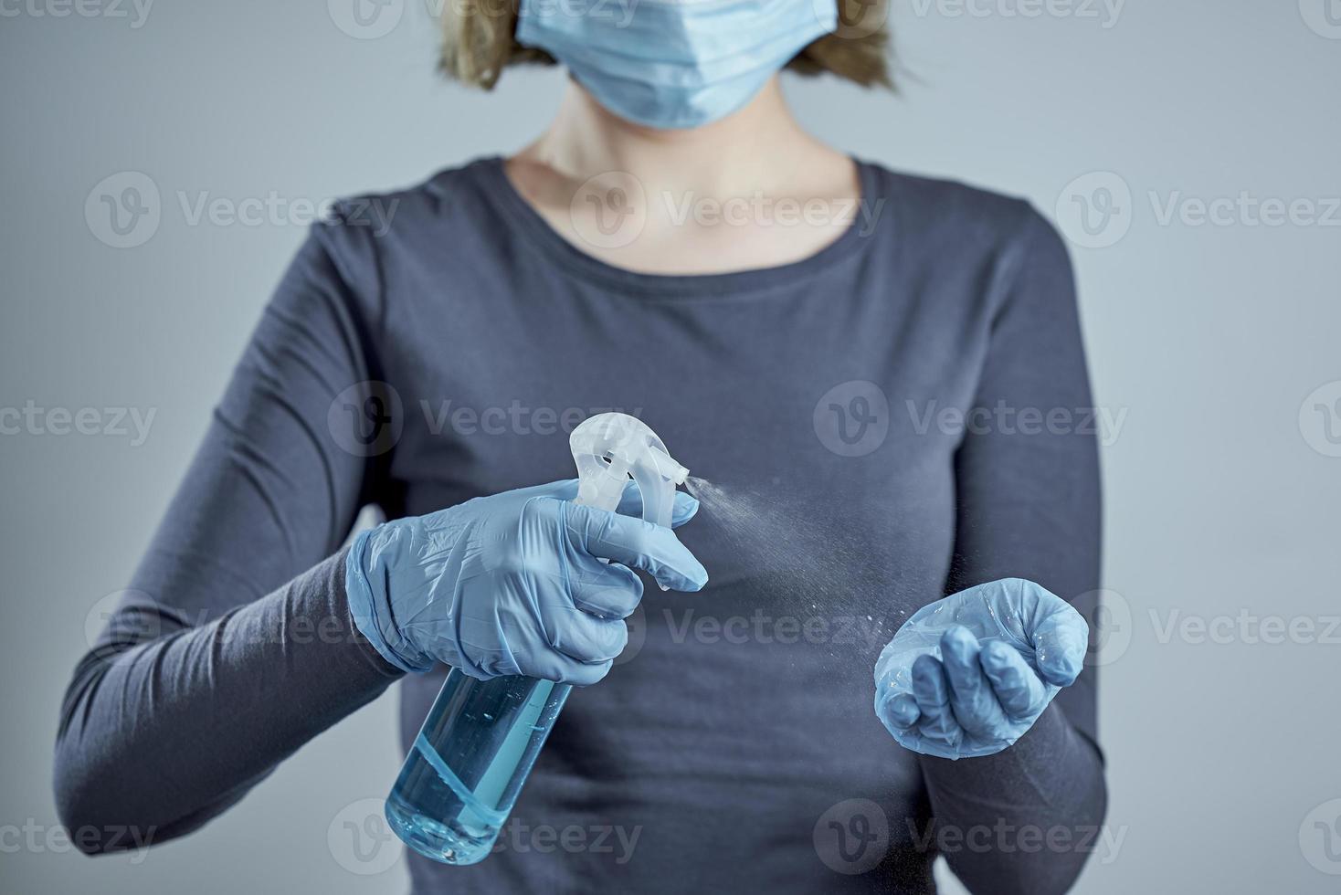 en flicka i en medicinsk mask behandlar sina händer i blåa engångshandskar med desinfektionsmedel på grå bakgrund. spray sanitizer på bakgrunden av en flicka foto