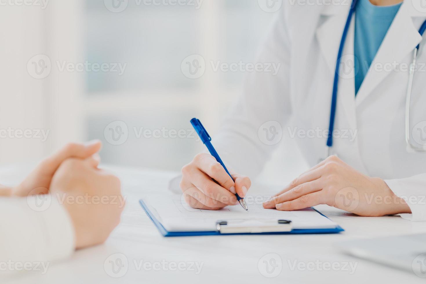 okänd kvinnlig läkare skriver ner recept på papper i urklipp, konsulterar patienten om att bota sjukdomar, poserar vid skrivbordet mot suddig vit bakgrund fyller upp medicinsk form. symtom diagnos foto