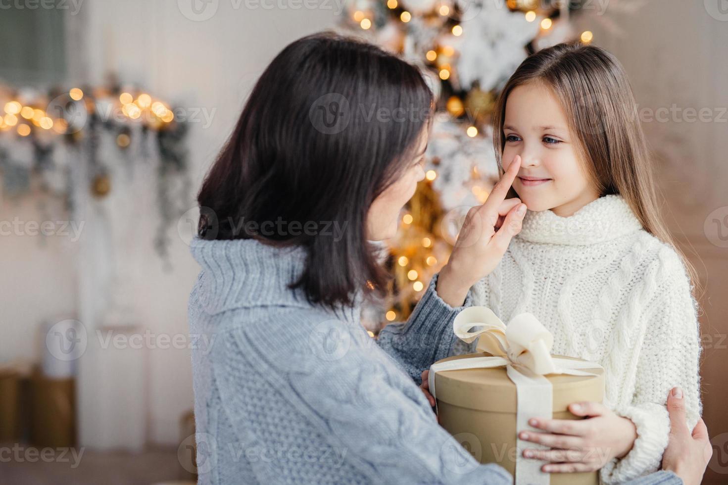 vackert kvinnligt barn med långt hår, bär vit varm tröja, ser ut i mammas ögon, glad över att få present på julen, fira vintersemester i familjekretsen. glad mor och dotter foto