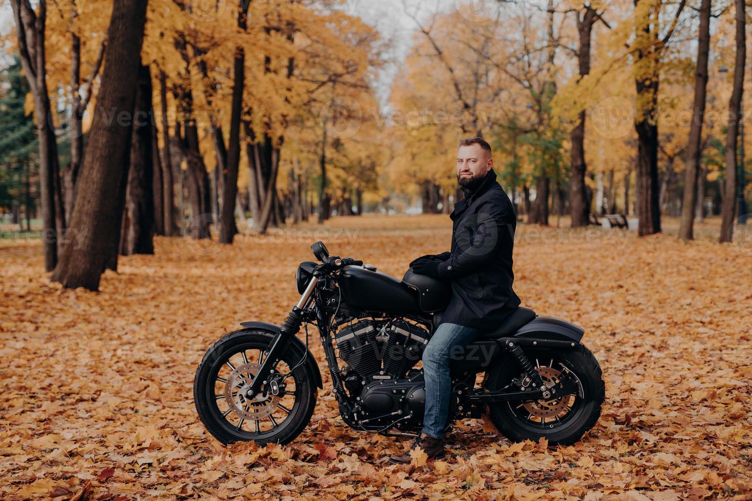 utomhusbild av manlig cyklist med tjockt skägg, bär skyddshandskar, svart kappa och jeans, poserar på motorcykel i vacker park med orange löv, håller hjälm, stannar för att vila. urban livsstil foto