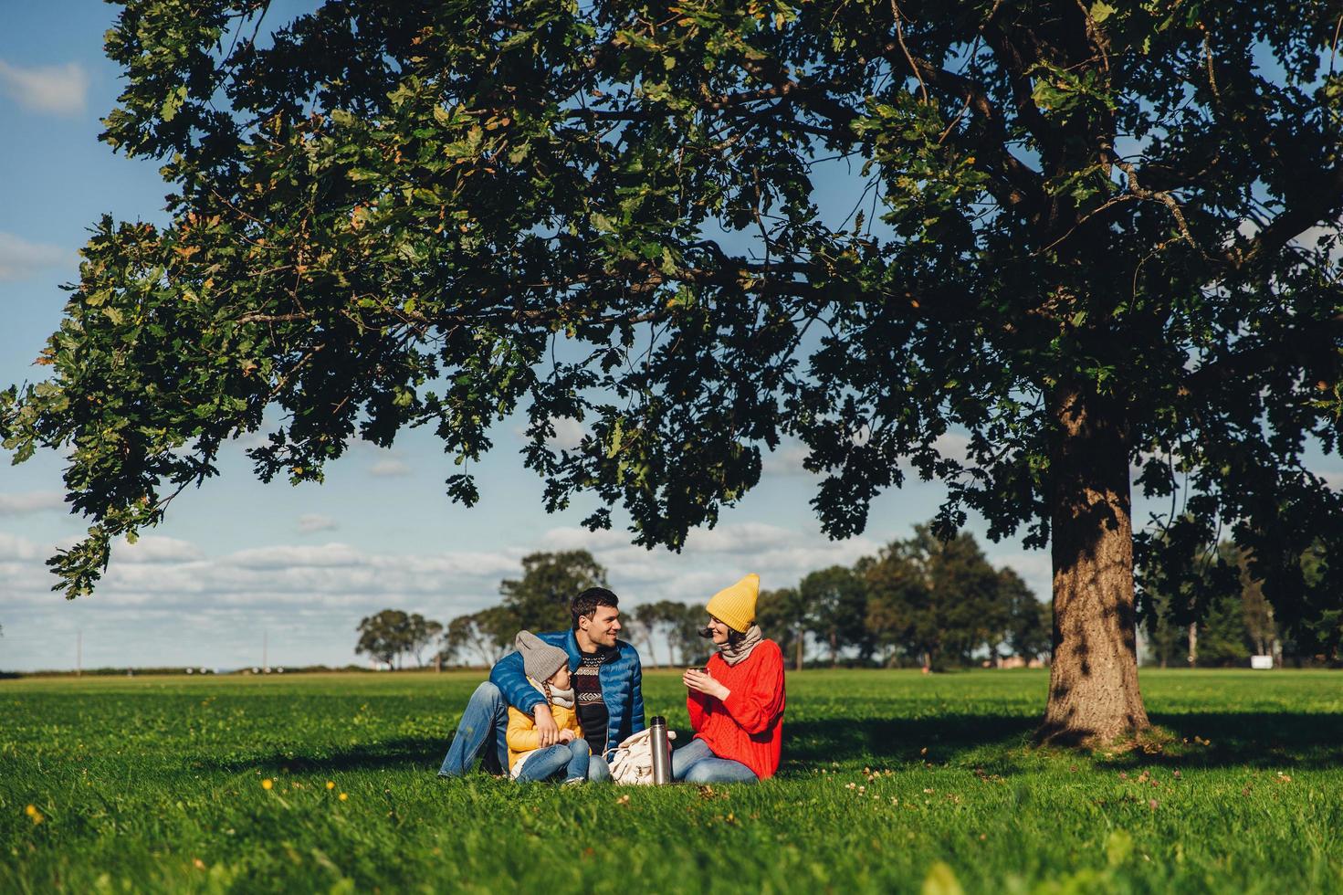 lycklig familj har höstpicknick, sitta på grönt gräs, dricka varmt te, kommunicera med varandra, njuta av lugn atmosfär, beundra blå himmel och vackra landskap. vänliga familjemedlemmar tillsammans foto