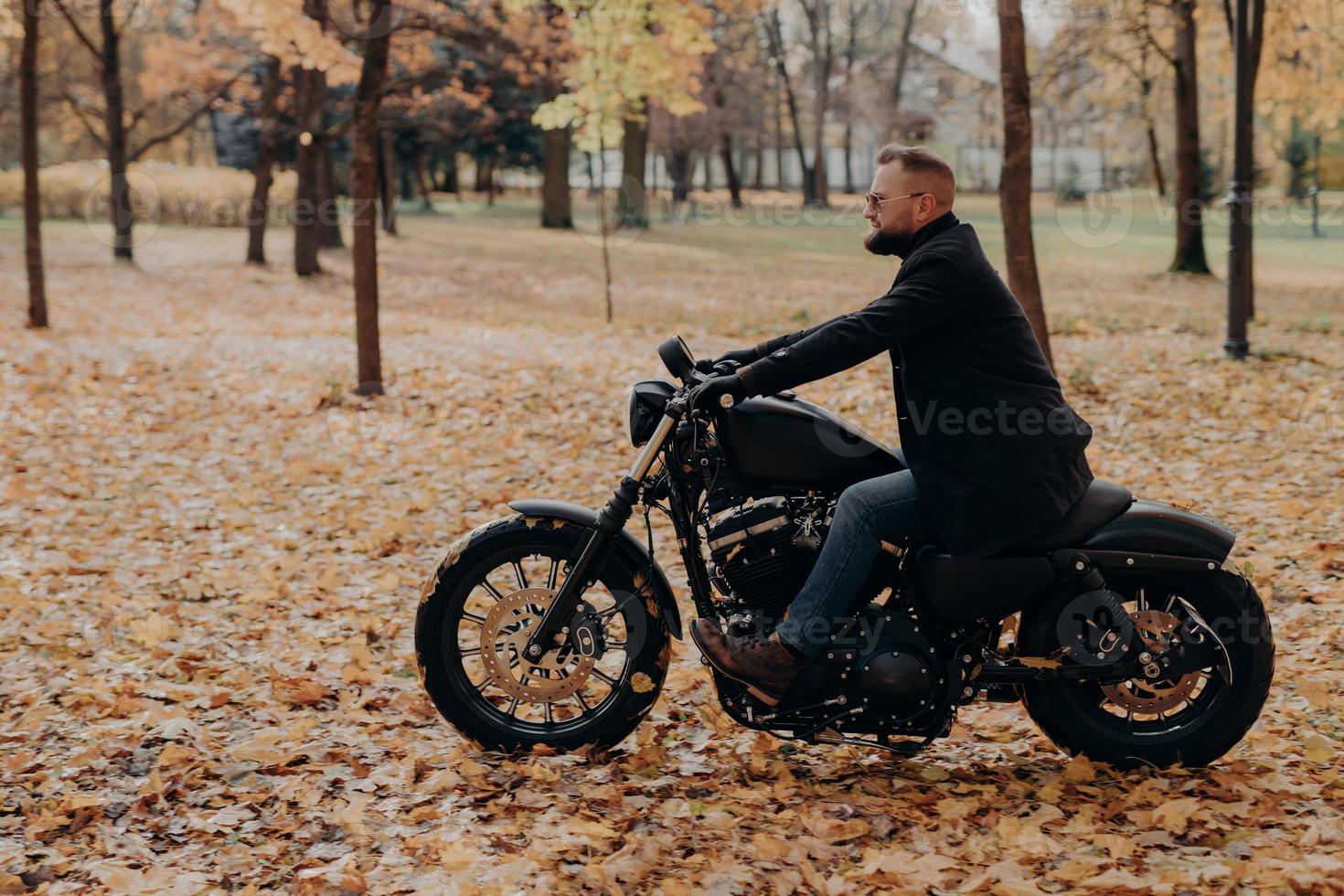 sidledsbild av självsäker manlig cyklist åker motorcykel, har äventyr på två hjul i höstparken, bär snygga kläder, skyddande solglasögon, njuter av naturen under den vackra höstsäsongen. foto