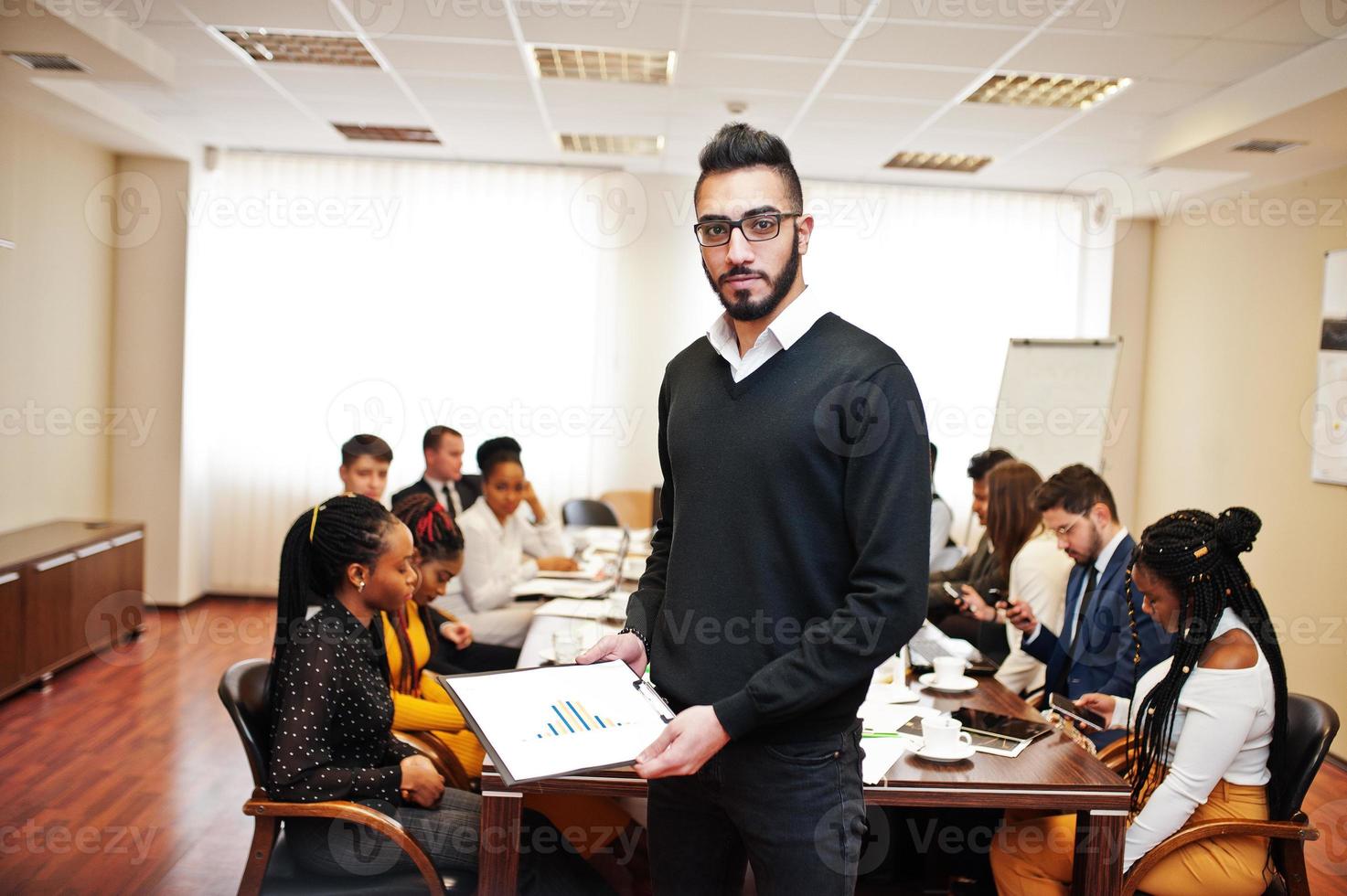 ansikte av stilig arabisk affärsman, håller urklipp på bakgrunden av affärsfolk multiracial team möte, sitter i kontorsbordet. foto