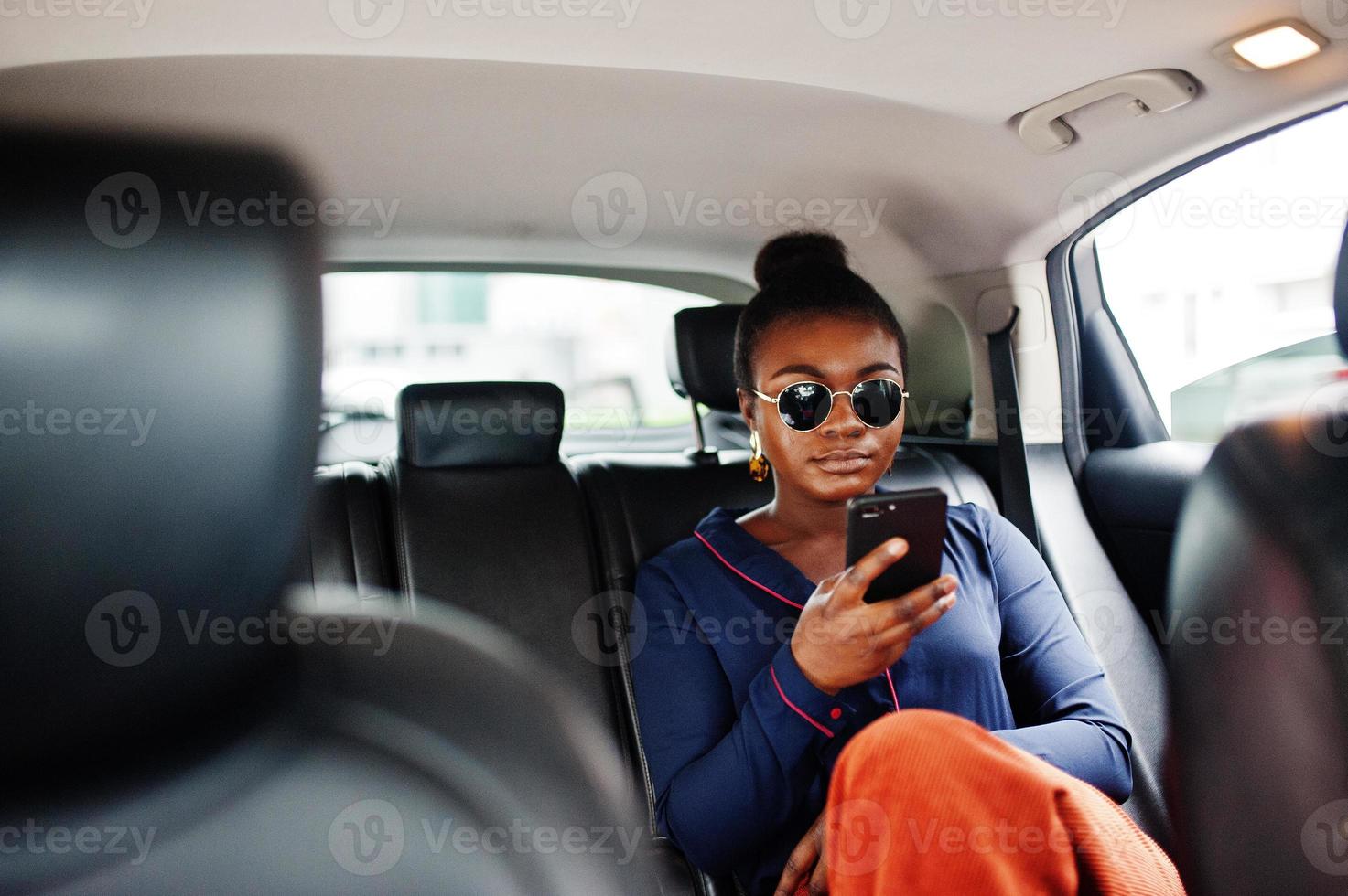 rik affärs afrikansk kvinna på solglasögon sitter vid suv bil med svarta lädersäten. mobiltelefon till hands. foto