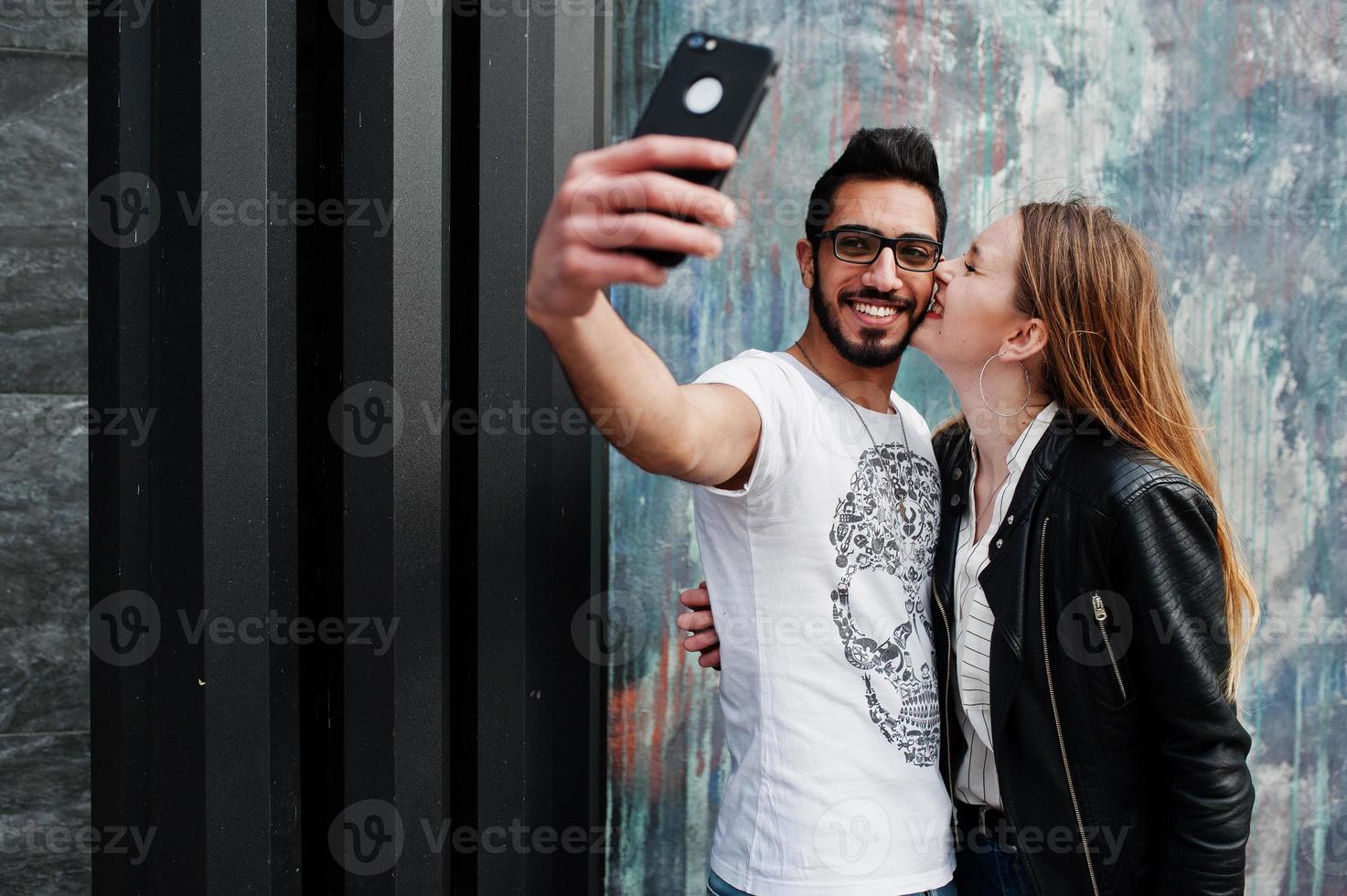 coola multiracial par poserar mot väggen och gör selfie tillsammans. europeisk flicka kysser arabisk man på kinden. foto