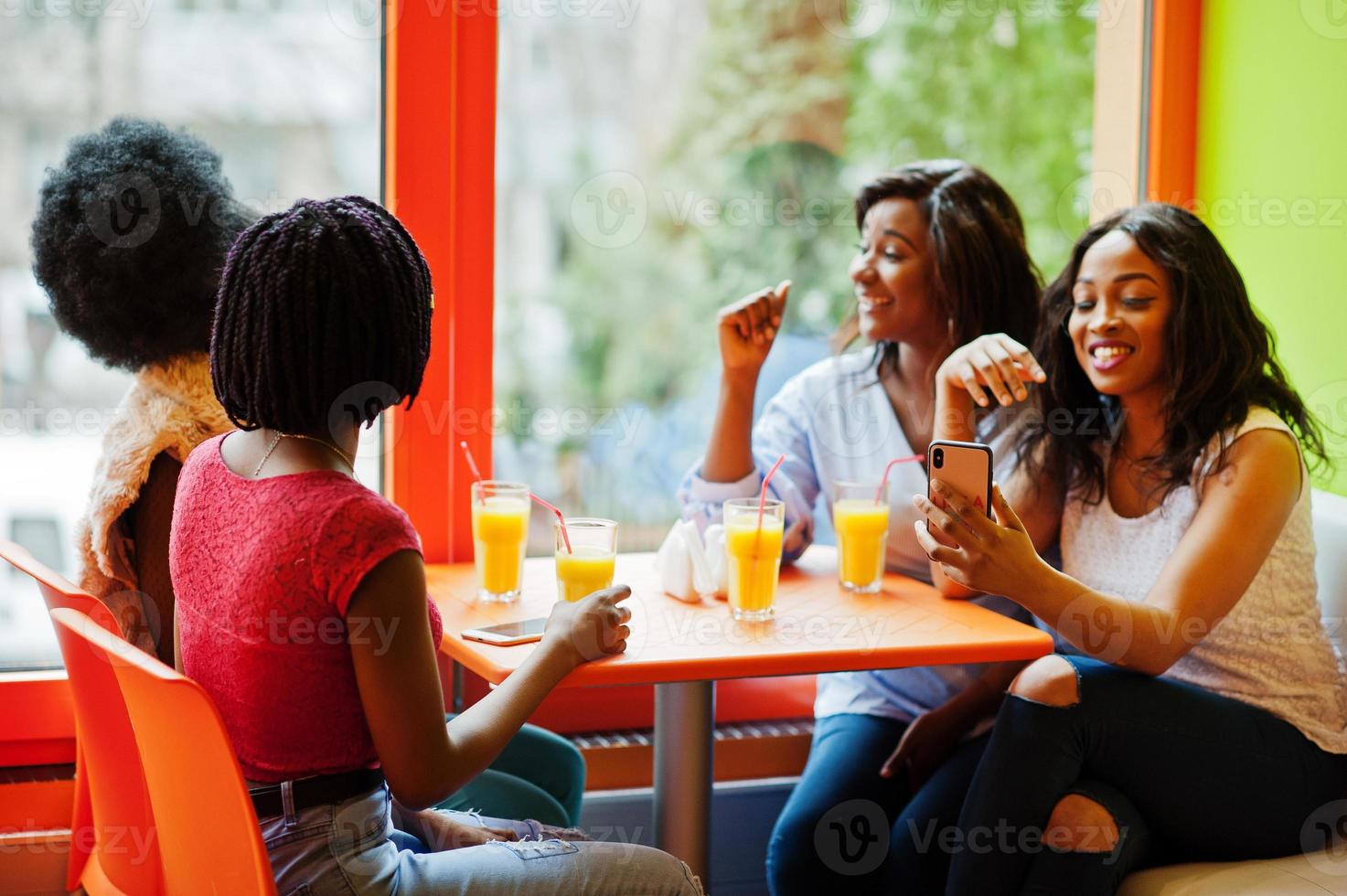 fyra unga afrikanska flickor med juice sitter i färgglada snabbmatsrestaurang och gör selfie via telefon. foto