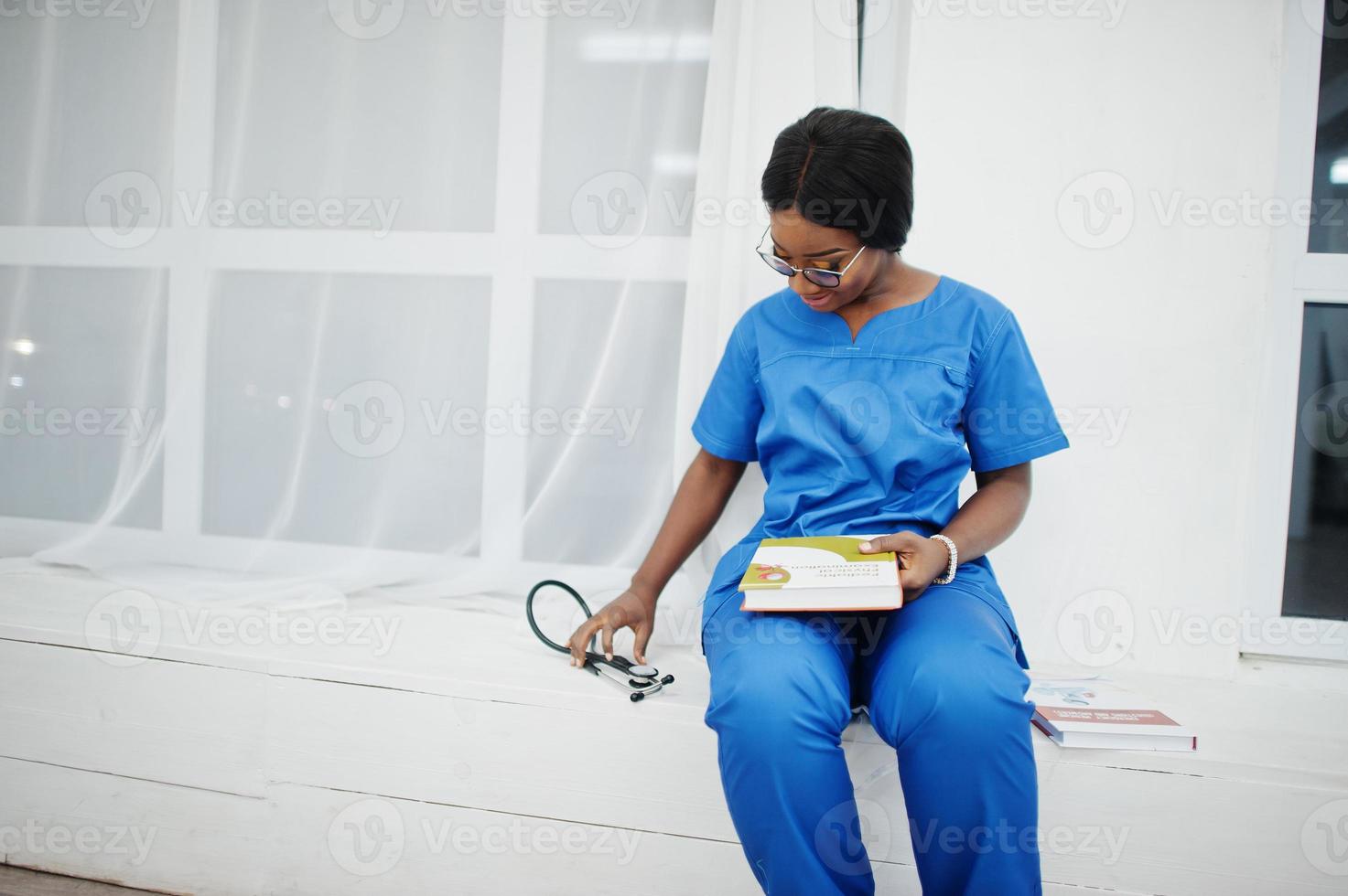 porträtt av glad kvinnlig afroamerikansk ung läkare barnläkare i blå uniform rock och stetoskop med böcker till hands. hälsovård, medicin, medicin specialist - koncept. foto