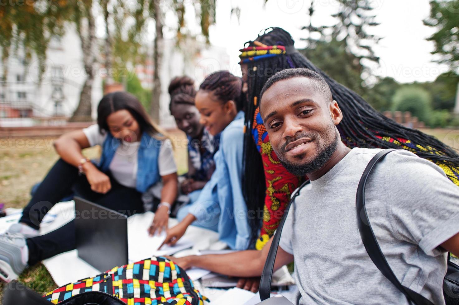 grupp på fem afrikanska högskolestudenter som spenderar tid tillsammans på campus på universitetsgården. svarta afro vänner sitter på gräs och studerar med bärbara datorer. foto