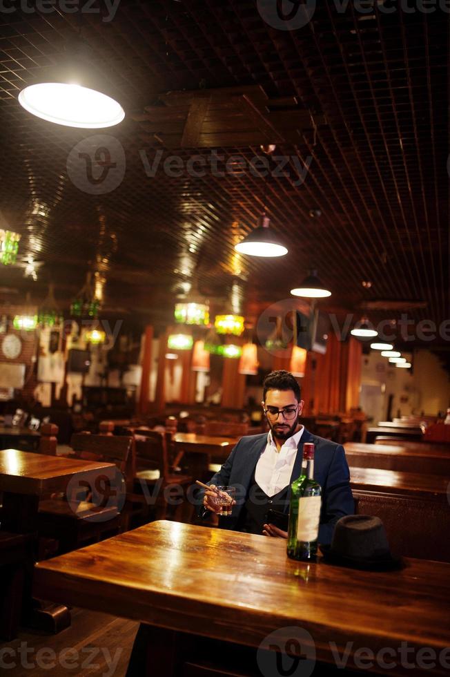 stilig välklädd arabisk man med ett glas whisky och cigarrhåll mobiltelefon, poserade på puben. foto