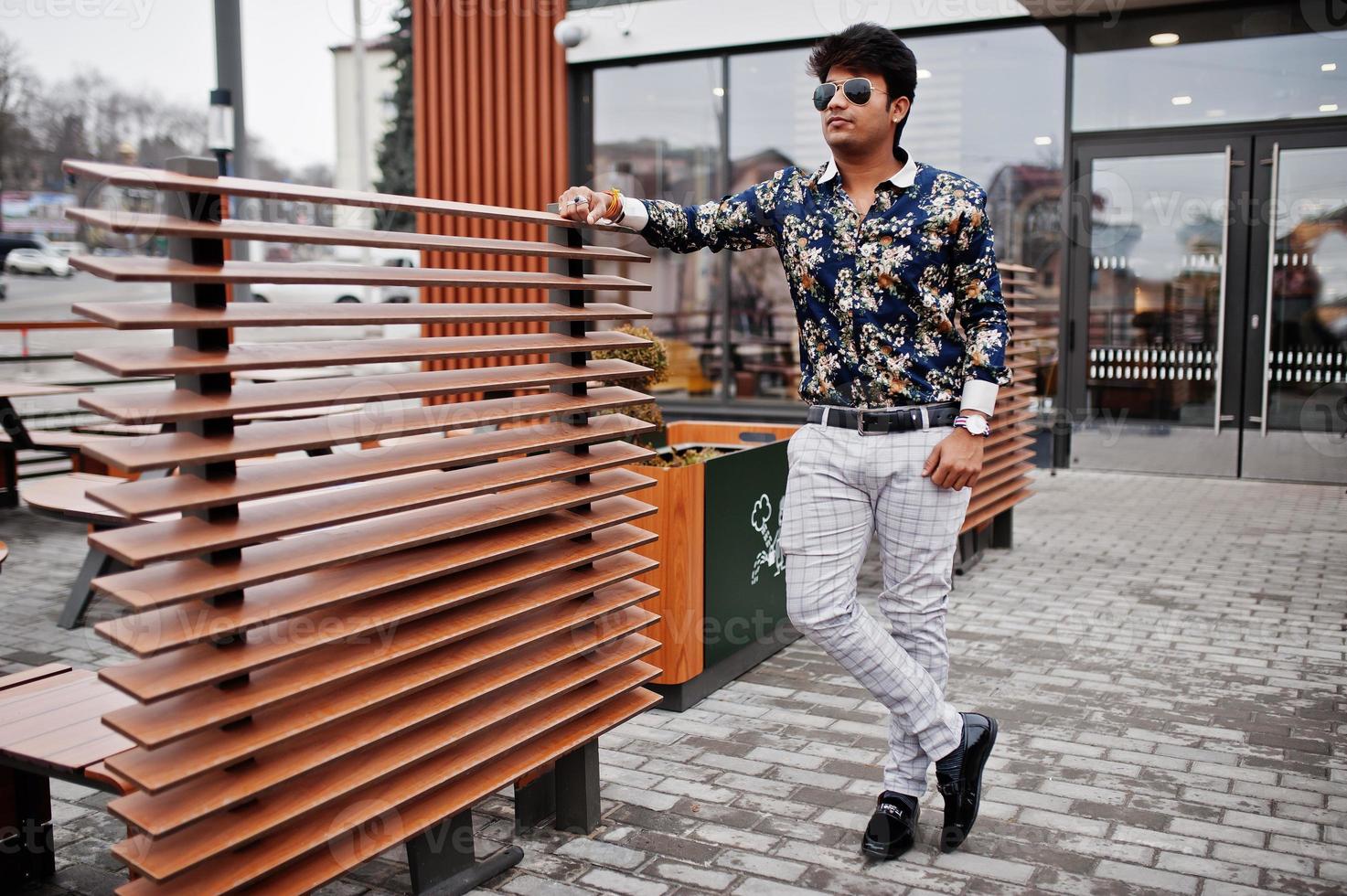 attraktiv och stilig snygg indisk man bär skjorta och solglasögon som står på terrassen till moderna loungecafé på sin arbetsrast. foto