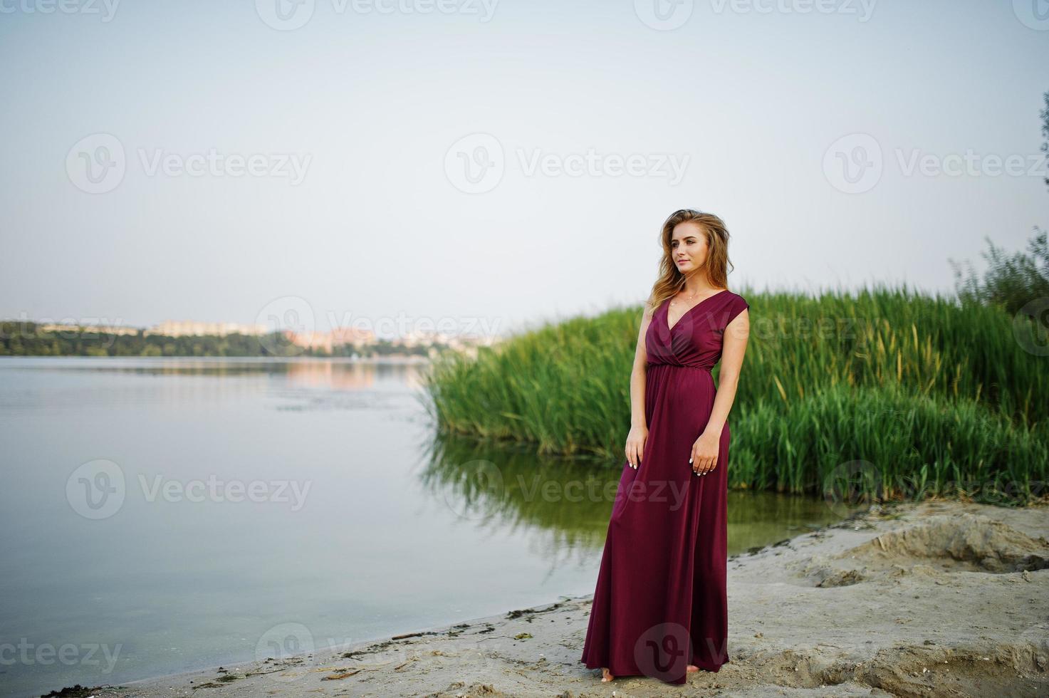 blond sensuell barfota kvinna i röd marsala klänning poserar mot sjön med vass. foto