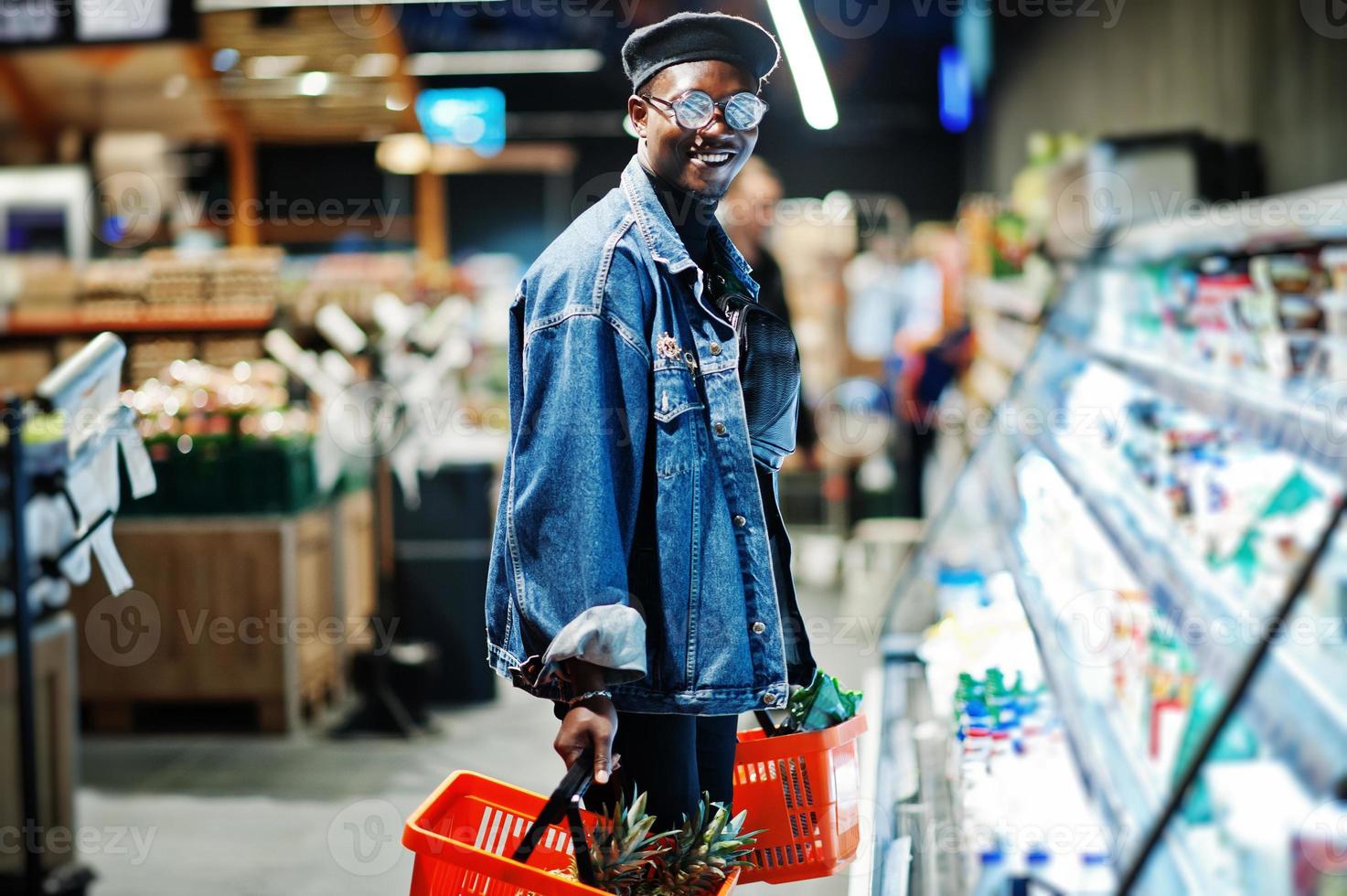 snygg casual afrikansk amerikansk man på jeansjacka och svart basker som håller två korgar, står nära kylskåpet och handlar i snabbköpet. foto