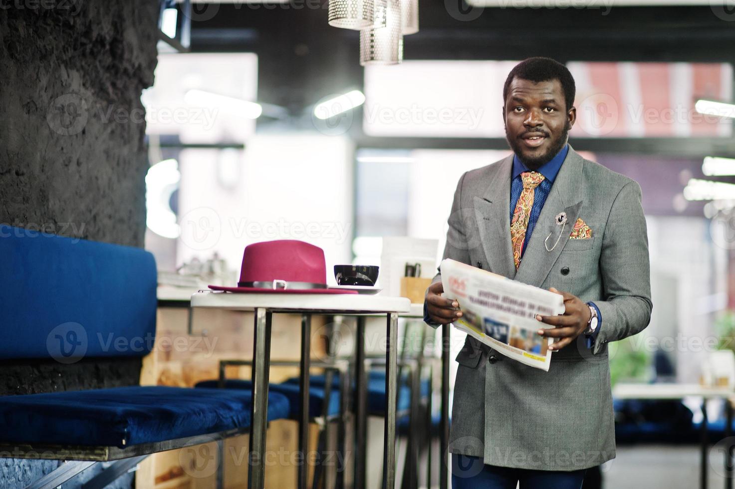 snygg afrikansk amerikansk man modell i grå jacka slips och röd hatt dricka kaffe på café och läsa tidningar. foto