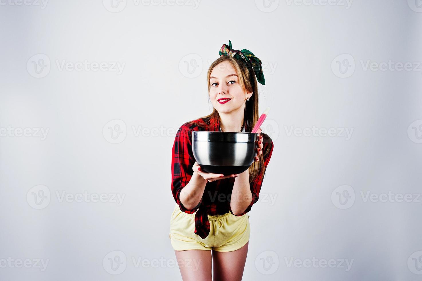 ung rolig hemmafru i rutig skjorta och gula shorts pin up stil med kastrull och kök sked isolerad på vit bakgrund. foto