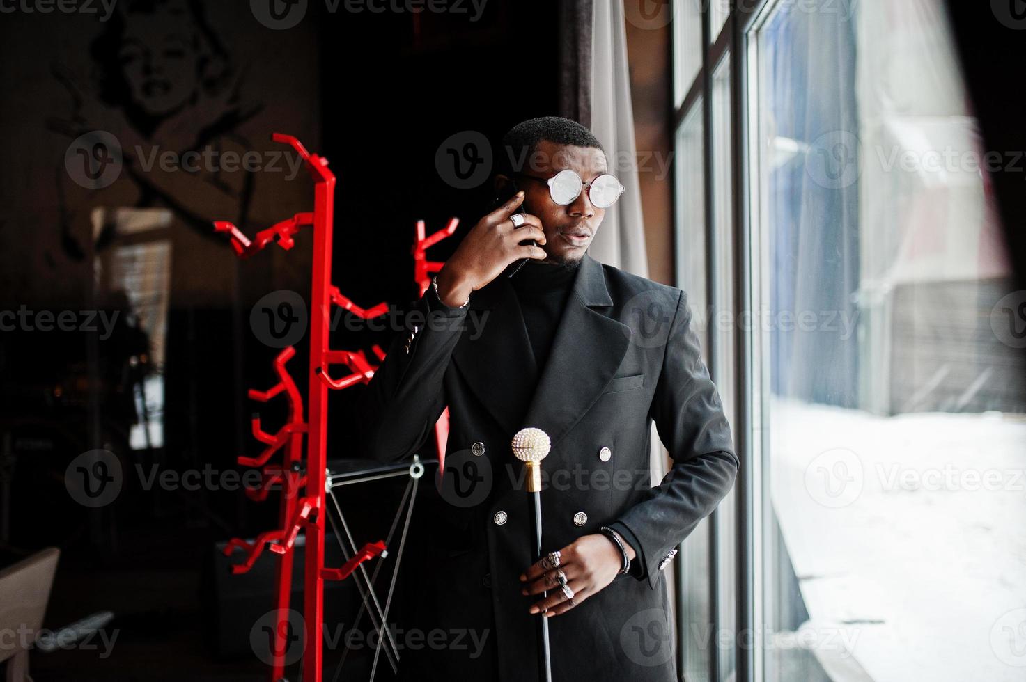 stilig afrikansk amerikansk herre i elegant svart jacka och glasögon, håller retro käpp som käppkolv eller tippande käpp med gyllene diamantkulhandtag och talar i mobiltelefon. foto