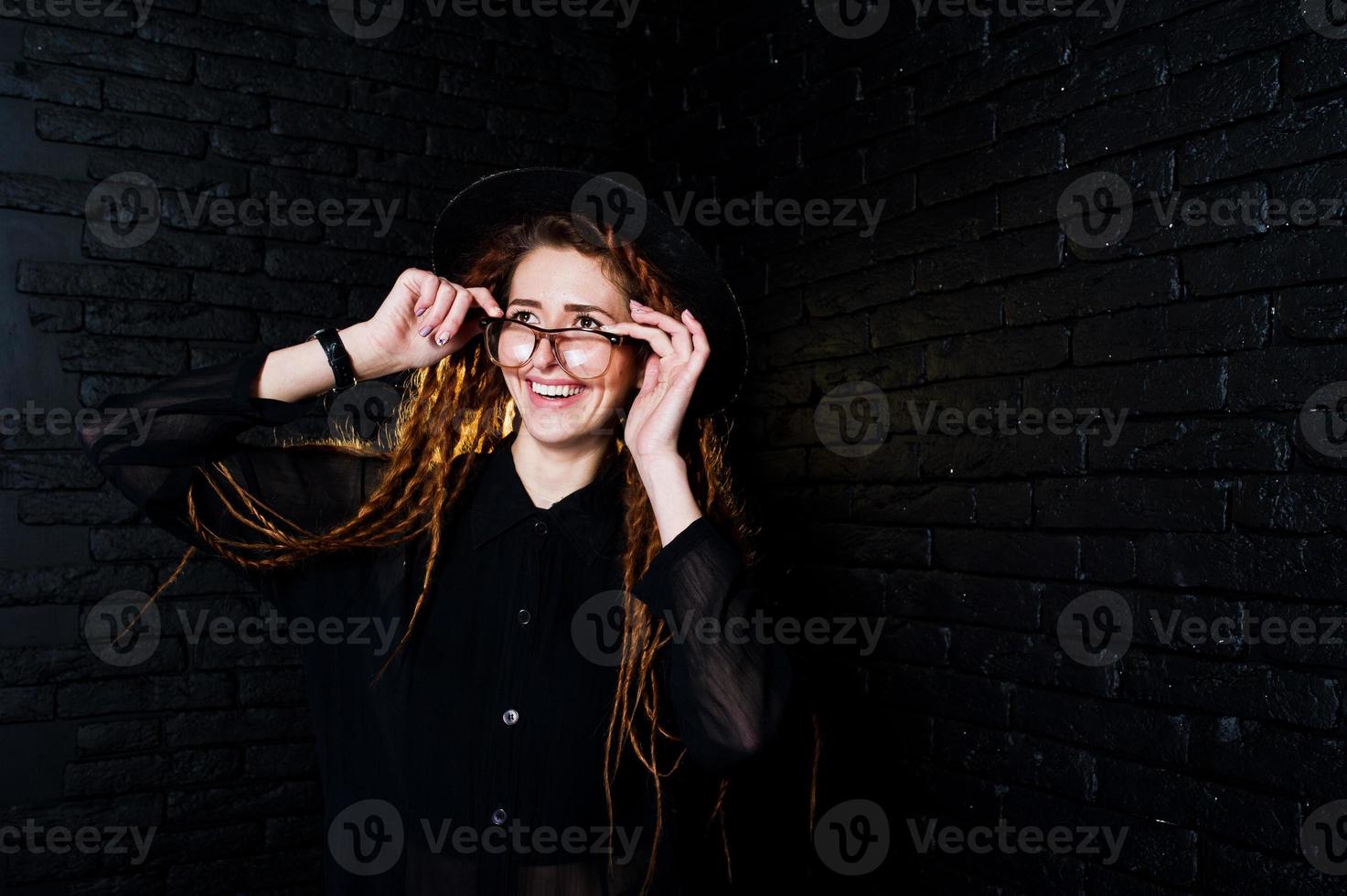 studiofotografering av flicka i svart med dreads, vid glasögon och hatt på tegelbakgrund. foto