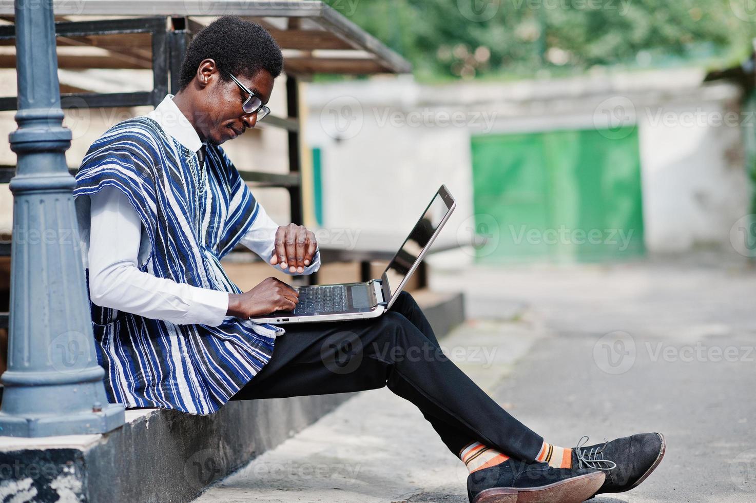 afrikansk man i traditionella kläder och glasögon med bärbar dator som arbetar utomhus och tittar på tiden för sina klockor. foto