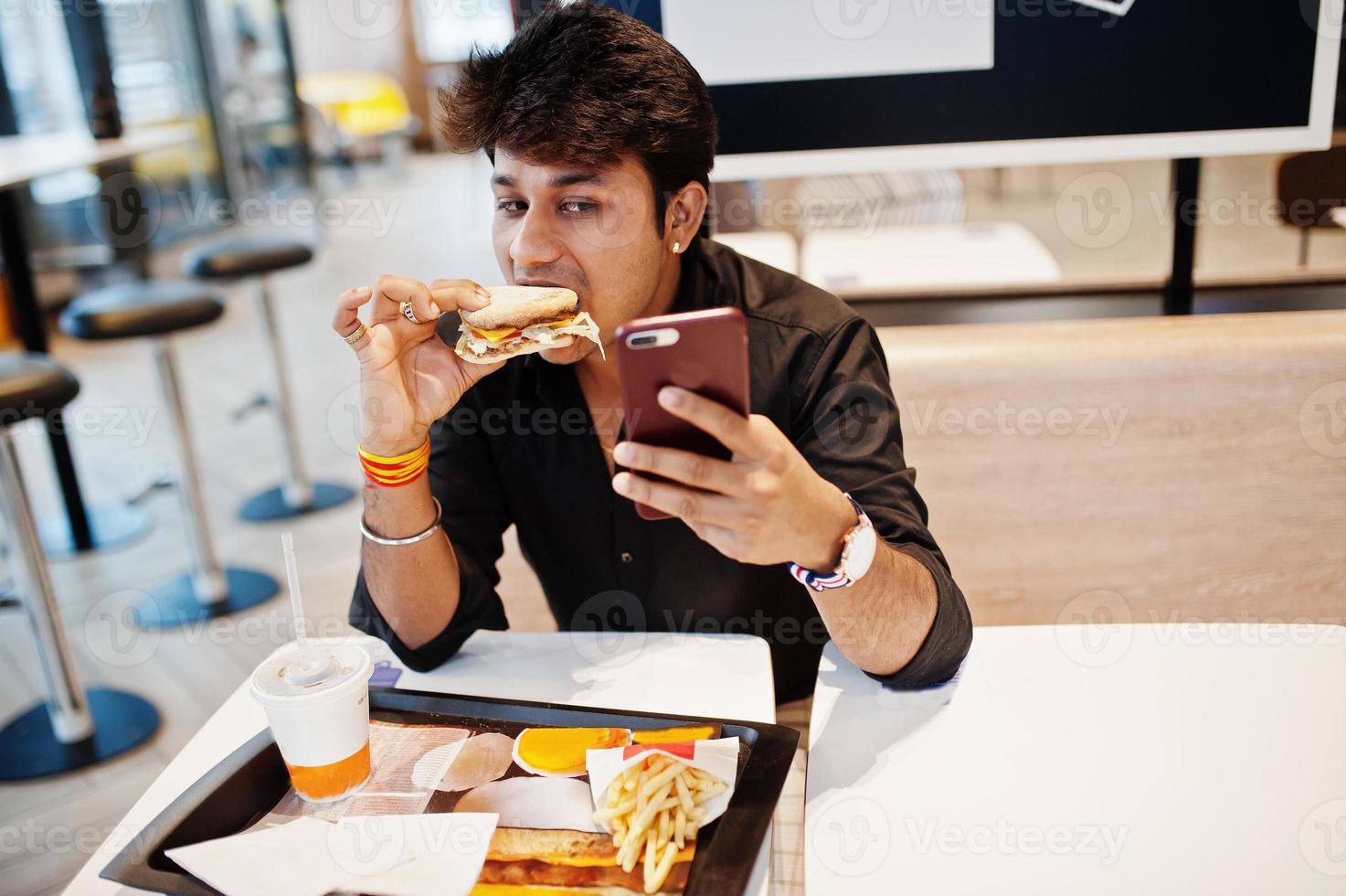 snygg och rolig indisk man sitter på snabbmatscafé och äter hamburgare och gör selfie via telefon. foto