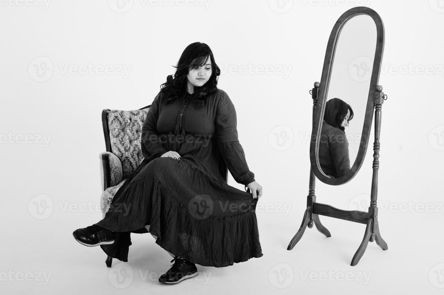 attraktiv sydasiatisk kvinna i djupröd klänning klänning poserade i studio på vit bakgrund mot spegel och sitter vid stolen. foto