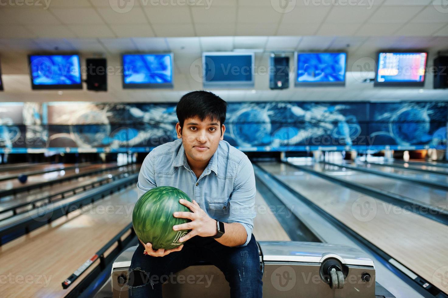 snygg asiatisk man i jeansskjorta stående vid bowlinghall med boll till hands. foto