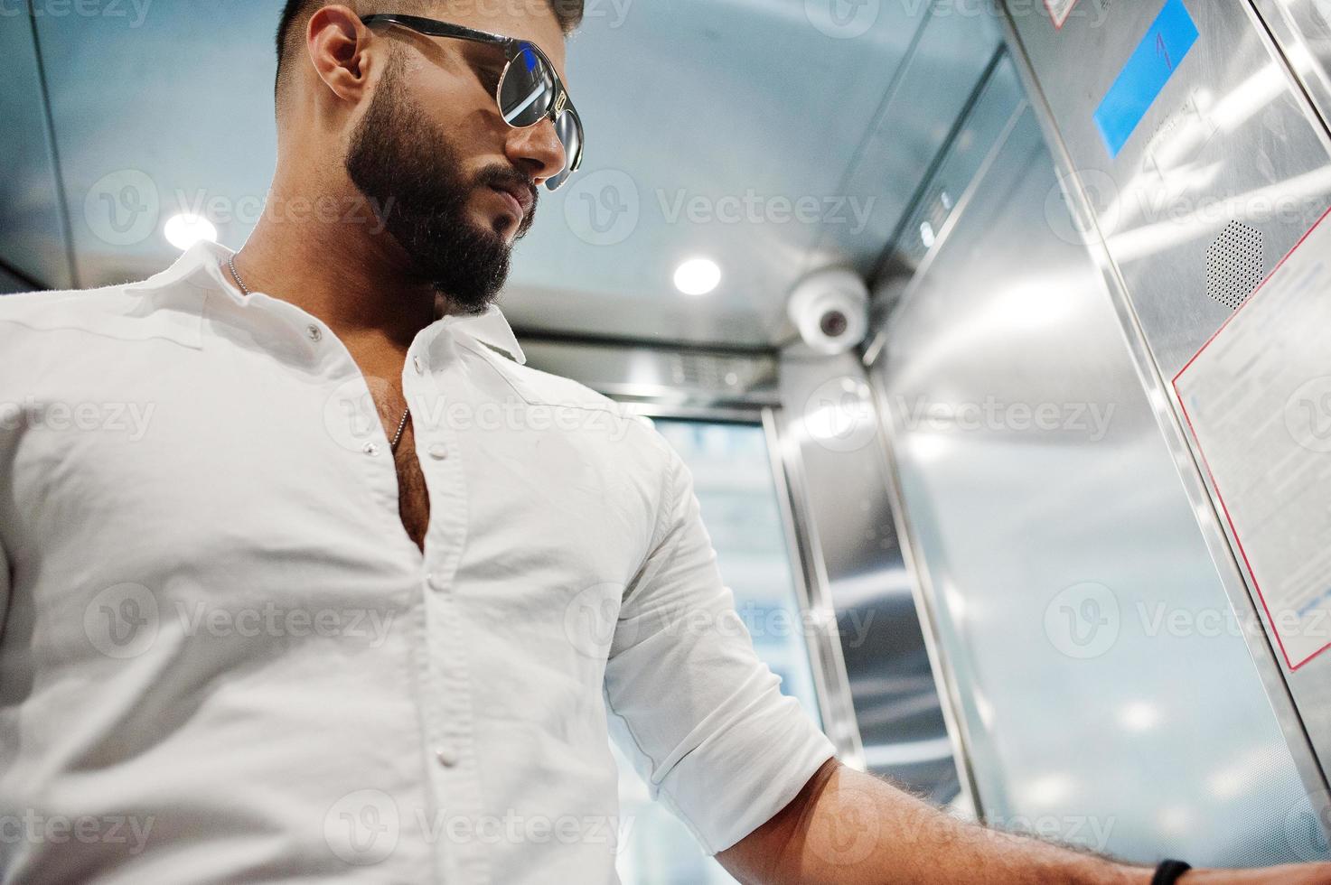 snygg lång arabisk man modell i vit skjorta och solglasögon poserade vid hissen inuti. foto