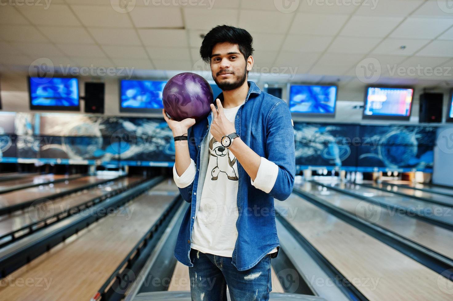 elegant skägg asiatisk man i jeansskjorta stående vid bowlinghall med boll till hands. foto