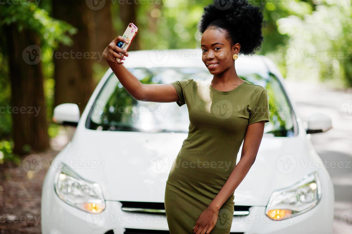afrikansk amerikansk kvinna poserade mot vit bil i skogsväg och tittar på mobiltelefonen. foto