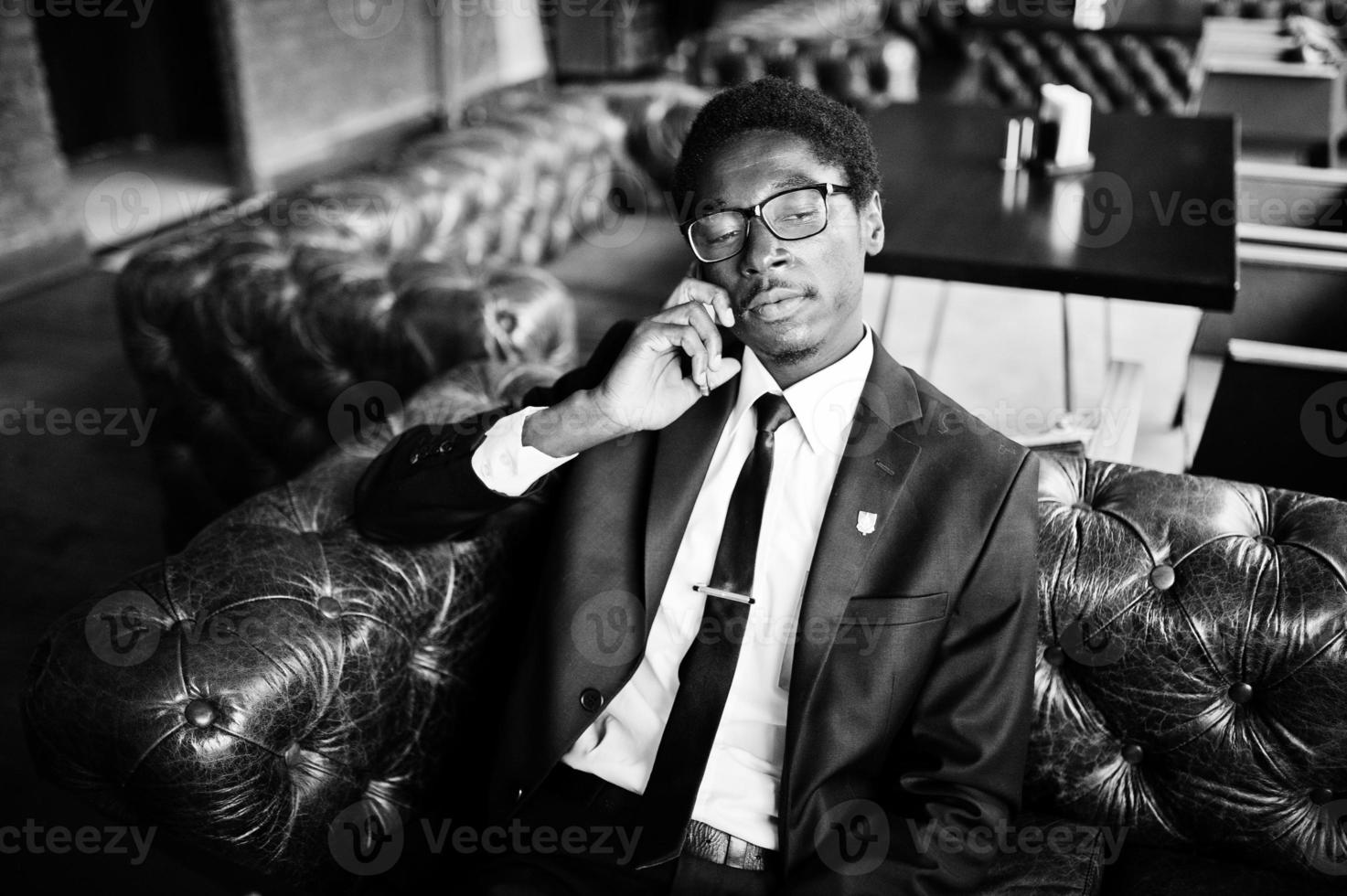 affärsafrikansk amerikansk man bär på svart kostym och glasögon sitter på kontoret och talar i telefon. foto