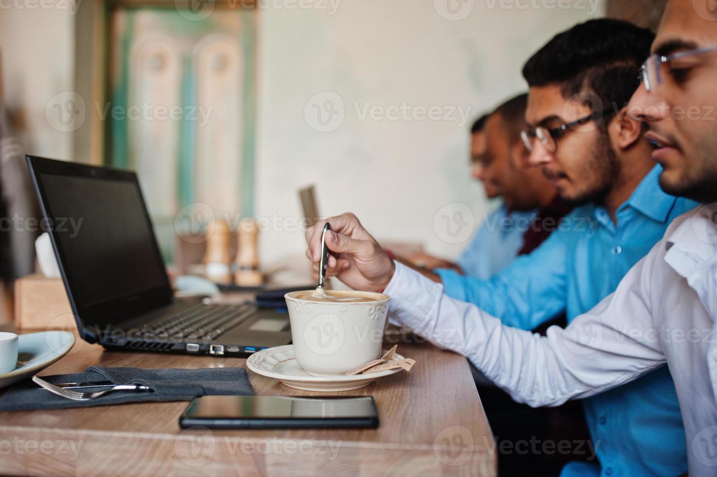 grupp av fyra sydasiatiska män poserade på affärsmöte i café. indianer arbetar med bärbara datorer tillsammans med olika prylar, pratar och dricker kaffe. foto