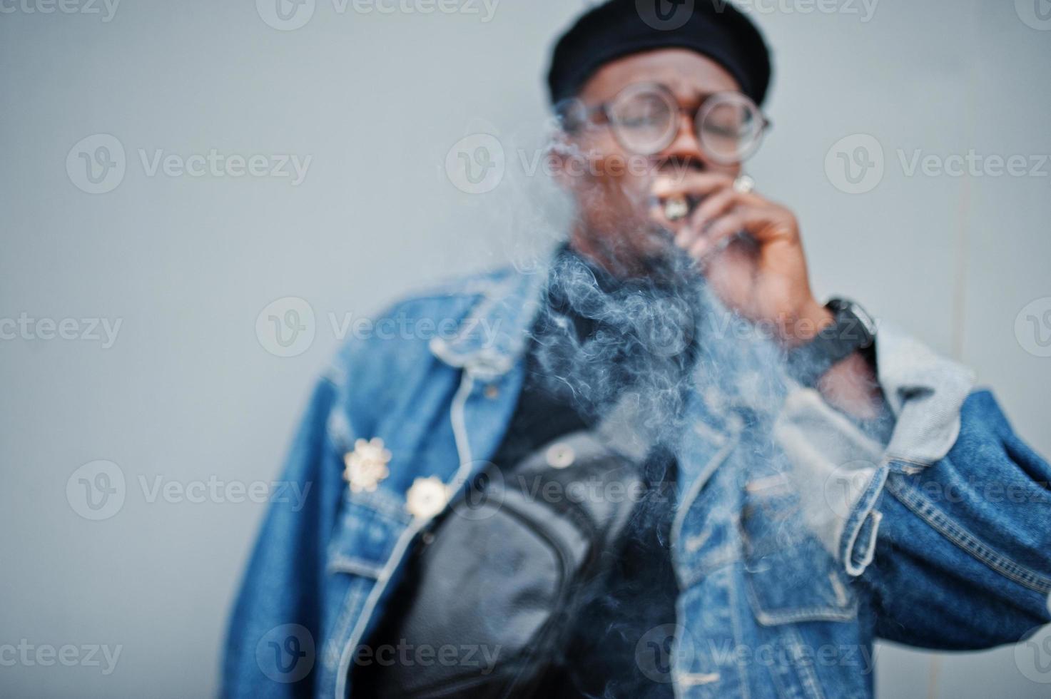 afrikansk amerikansk man i jeansjacka, basker och glasögon, röker cigarr på grå stålbakgrund. foto