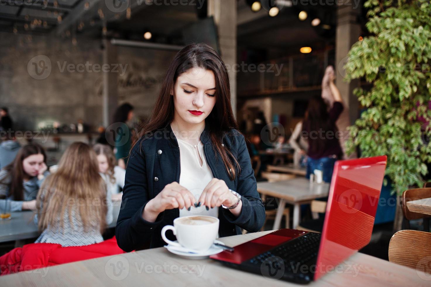 brunett flicka sitter på café med kopp cappuccino, arbetar med röd laptop. foto